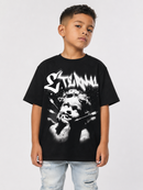 A young boy wearing a black graphic t-shirt with a white stylized logo and a high-contrast portrait print, paired with light-wash distressed jeans, photographed against a light gray studio background.