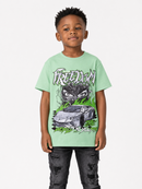 A young boy wearing a light green graphic t-shirt with a black and white car illustration and the word 'FREEDOM' on the front, paired with distressed black jeans, standing against a light gray background.