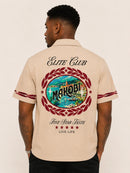 A man viewed from the back wearing a beige short-sleeve camp collar shirt with a large graphic print on the back. The print features the words 'ELITE CLUB' and 'Five Star Taste LIVE LIFE' with a central circular design showing a scenic lake and the word 'MAKOBI' inside a red laurel wreath. By Fresh N Fitted