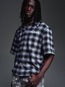 A man wearing a black and white plaid short-sleeve button-up shirt with 'GUAPI' printed in white on the chest, posed against a dark gray studio background. By Fresh N Fitted.