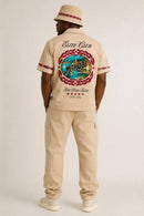 A man shown from the back wearing a beige short-sleeve button-up shirt with a large circular Morocco graphic and 'Elite Club' text, paired with matching beige cargo pants and a beige bucket hat with red trim, against a light gray studio background. By Fresh N Fitted