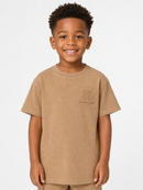 A young boy smiling and wearing a light brown short-sleeve t-shirt with a tonal embroidered logo on the left chest, set against a plain light gray background.
