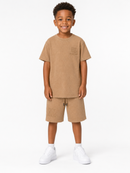 A young boy stands against a plain white studio background wearing a matching tan short-sleeve t-shirt and shorts set with white sneakers.