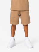 A child wearing beige drawstring shorts with an embossed logo on the left leg, styled with a matching beige top and white sneakers against a light gray studio background.