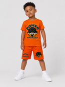 A young boy modeling a bright orange two-piece athletic set consisting of a short-sleeve t-shirt and matching shorts, styled with white sneakers against a light gray studio background.