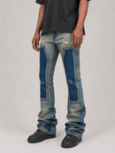 A person wearing light-wash distressed denim jeans with large blue patch panels on the thighs and knees, styled with a dark t-shirt and black shoes against a light gray studio background.