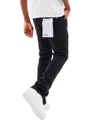 A person wearing black slim-fit jeans with a white patterned rectangular patch pocket on the back, paired with a white t-shirt and white sneakers, shown against a plain white background.