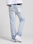 A person wearing light-wash blue distressed jeans with paint-splatter details and a white patterned paper patch in the back pocket, paired with a white t-shirt and white sneakers against a light gray studio background.
