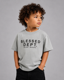 A young child with curly brown hair wearing a light gray crewneck t-shirt with 'BLESSED DEPT. BEYOND MEASURE' printed in black on the front, paired with black pants, against a solid gray studio background.