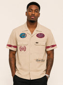 A man wearing a beige short-sleeve button-up shirt with red stripe details on the sleeves, embroidered patches on the chest, and 'Sun Club' text on the lower front, paired with black pants against a light beige background. By Fresh N Fitted