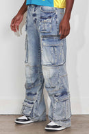 A person wearing very baggy light-wash blue denim cargo pants with multiple flap pockets and distressed details, paired with black and white sneakers against a plain indoor wall. By Fresh N Fitted.