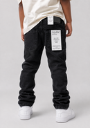 A child shown from the back wearing black denim jeans with white paper labels on the back pockets, a cream t-shirt, and white sneakers, photographed against a light gray studio background.