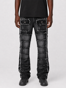 A person wearing heavily distressed black denim jeans with raw frayed edges and patchwork paneling, paired with a black t-shirt and black shoes, against a light gray studio background.