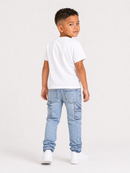 A young boy standing in profile, wearing a plain white short-sleeve t-shirt, light blue cargo-style jeans, and white sneakers against a clean white studio background.