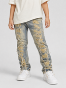 A child wearing distressed light blue denim jeans with heavy gold and white thread embroidery and frayed details, styled with a cream t-shirt and white sneakers against a light gray studio background.