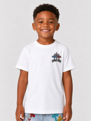 A young boy smiling and wearing a white short-sleeve crewneck t-shirt with a small multicolor cross graphic on the left chest, set against a plain light gray background.