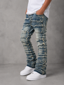 A person wearing heavily distressed and frayed blue denim jeans with a baggy, ruffled fit, styled with a white t-shirt and white sneakers against a gray concrete background.