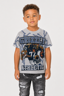 A young boy wearing a light gray t-shirt with a 'Grizzly Athletic' grizzly bear graphic and distressed black jeans, standing against a light gray background.