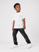 A young boy modeling a white graphic t-shirt, distressed black jeans, and white sneakers against a plain light gray studio background.