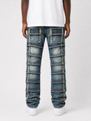 Rear view of a person wearing blue patchwork denim jeans with frayed trim and multiple stitched panel details, paired with a white t-shirt and white sneakers on a light gray studio background.