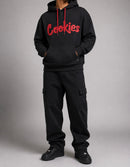 A model wearing a black pullover hoodie with a red 'Cookies' logo on the chest, paired with black cargo pants and black sneakers, against a neutral gray studio background. By Fresh N Fitted