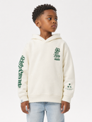 A young boy wearing a cream-colored hoodie with green graphics and light blue jeans, standing against a plain white studio background.