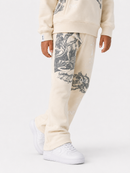 A child wearing cream-colored sweatpants with large black graphic prints on the legs, paired with a matching cream sweatshirt and white sneakers, shown against a light gray studio background.