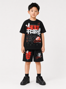 A young boy modeling a black graphic t-shirt and matching black shorts with red and white print details, paired with black socks and black sneakers, against a light gray studio background.