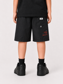 A child shown from the back wearing black shorts with small red and white printed graphics, a black t-shirt, black socks, and black sneakers, against a light gray studio background.