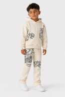 A young boy modeling a cream-colored two-piece tracksuit with dark gray graphic prints, consisting of a pullover hoodie and matching sweatpants, paired with white sneakers on a light gray studio background.