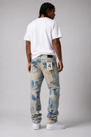A man viewed from behind wearing distressed light blue jeans with blue paint-splatter details, a plain white t-shirt, and white sneakers against a light gray studio background. By Fresh N Fitted