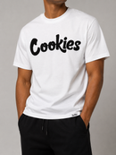 A white short-sleeve crewneck t-shirt with the word 'Cookies' printed across the chest in a bold black script font. The shirt is modeled by a person wearing black drawstring sweatpants against a neutral gray studio background. By Fresh N Fitted
