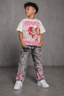 A young boy stands against a gray concrete wall wearing a beige graphic t-shirt with pink text and a cupid character print, paired with gray acid-wash jeans featuring pink flame and graphic prints, and white sneakers. By Fresh N Fitted.