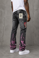 A person shown from the back wearing washed black denim jeans with pink flame graphics near the cuffs, a red skull patch on the upper leg, and a white t-shirt and white sneakers, standing against a gray concrete wall. By Fresh N Fitted.