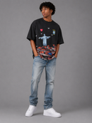 A man modeling a black graphic t-shirt with a multicolor print of Christ the Redeemer and Rio de Janeiro landmarks, paired with light-wash blue jeans and white sneakers, against a solid gray studio background. By Fresh N Fitted
