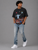 A man modeling a black graphic t-shirt with a vibrant Brazil-themed print, paired with light-wash blue jeans and white sneakers, against a solid gray studio background. By Fresh N Fitted