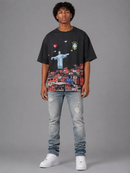 A man modeling a black graphic t-shirt with a vibrant print of Rio de Janeiro, paired with light-wash distressed jeans and white sneakers, against a solid gray studio background. By Fresh N Fitted