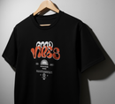 A black short-sleeve t-shirt hanging on a wooden hanger against a gray wall. The shirt features a retro-style graphic on the chest with the word 'VIBE' in wavy orange-red letters, layered over white text that reads 'GOOD VIBE'. By Fresh N Fitted.