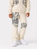 A child wearing cream-colored sweatpants with large black graphic prints of skulls and figures, paired with a matching cream sweatshirt and white sneakers, shown against a light gray studio background.