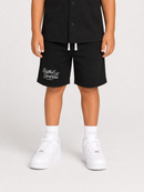 A child wearing black shorts with a white script logo on the left leg, a black short-sleeve button-up shirt, white socks, and white sneakers, shown from the waist down on a light gray studio background.