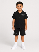 A young boy modeling a black short-sleeve button-up shirt and matching black shorts set, featuring white embroidered script logos on the chest and left leg, styled with white socks and white sneakers against a light gray studio background.