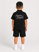 A young boy viewed from the back wearing a black short-sleeve polo shirt and matching black shorts. The back of the polo features white cursive text reading 'Destined For Greatness'.