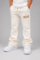 A person wearing cream sweatpants with a gold 'SEE YOU IN PARADISE' text graphic on the left thigh, styled with a white t-shirt and white sneakers against a neutral gray background.