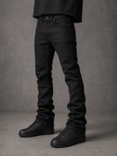 A person wearing slim-fit black jeans and black low-top sneakers, standing against a textured gray studio backdrop.
