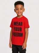 A young boy wearing a bright red crewneck t-shirt with the words 'WEAR YOUR FREEDOM' printed in distressed black block letters, paired with black pants, against a light gray studio background.