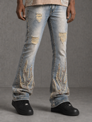Light-wash distressed denim jeans with ripped details and large embroidered flame motifs on the lower legs, shown on a model wearing black sneakers against a gray studio background.