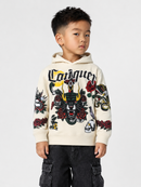 A young boy wearing a cream-colored Conquours hoodie with a large black, red, and yellow graphic print on the front and sleeves, paired with dark cargo pants, photographed against a light gray studio background.