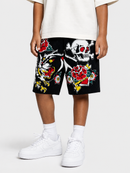 A person wearing black embroidered shorts with a tiger, skull, and red rose patchwork design, styled with a white t-shirt, white socks, and white sneakers against a light gray background.