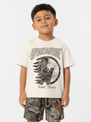 A young boy wearing a cream-colored t-shirt with a camouflage graphic print that says 'PARADISE Rebel Minds' and matching camouflage shorts, standing against a light gray studio background.