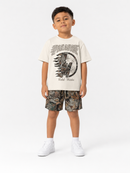 A young boy stands against a light gray studio background wearing a cream graphic T-shirt, camouflage shorts, and white sneakers. The T-shirt features a large distressed graphic with the word 'PARADISE' and smaller text below.
